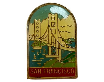 Op de afbeelding: Een gouden pin met een afbeelding van de Golden Gate Bridge in San Francisco. De brug is geel en de achtergrond is blauw en groen. De tekst "SAN FRANCISCO" staat onderaan de pin.