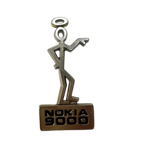 Op de afbeelding: Een zilverkleurige metalen speld in de vorm van een gestileerd stokfiguurtje met een halo boven het hoofd, staand op een rechthoekige basis. Op de basis staat de tekst NOKIA 9000 in zwarte letters afgebeeld. Het figuurtje heeft één arm uitgestrekt.