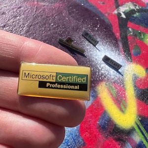May include: A Microsoft Certified Professional pin with a yellow background and green text. Also, three small black and gray pins with the word "Start" on them. The items are on a colorful, graffiti-covered surface.
