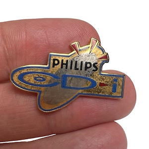 May include: Vintage Philips CD-i pin. The pin features a gold-colored outline with blue and red accents. The text "PHILIPS CD-i" is prominently displayed in black. The pin is a collectible item, likely from the 1990s.