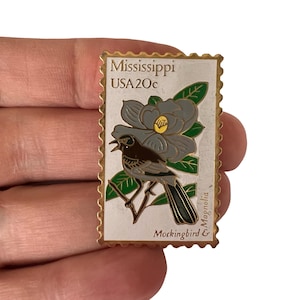 Op de afbeelding: Rechthoekige speld met een ontwerp dat doet denken aan een vintage postzegel. Het toont een grijze vogel op een tak, met een grijze magnoliabloem. Tekst bevat "Mississippi USA 20c" en "Mockingbird & Magnolia".