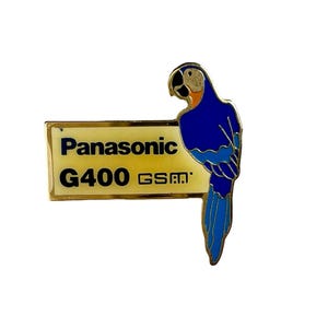 Puede incluir: Un pin de color dorado con un loro azul y naranja posado sobre una pancarta amarilla. La pancarta dice "Panasonic G400 GSM".
