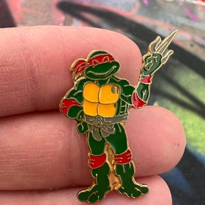 May include: A colorful enamel pin featuring the Teenage Mutant Ninja Turtle Raphael. The pin is green, red, yellow, and gold, with Raphael in a dynamic pose. The pin is held in a person's hand.