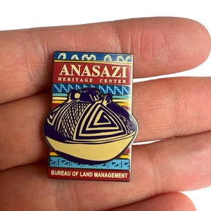 May include: A rectangular pin featuring the Anasazi Heritage Center logo. The pin has a red and blue background with the words "ANASAZI HERITAGE CENTER" in white. A stylized pot is in the center, with the words "BUREAU OF LAND MANAGEMENT" at the bottom.