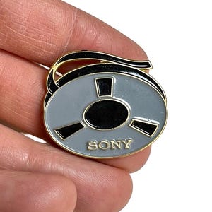 Könnte beinhalten: Ein grauer und schwarzer Sony-Tonbandstift mit goldenem Rand. Der Stift trägt das Wort "SONY" in goldfarbenen Buchstaben. Der Stift ist ein Accessoire im Vintage-Stil.
