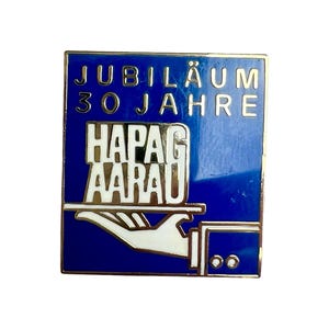 May include: A blue and gold enamel pin with the text "JUBILÄUM 30 JAHRE HAPAG AARAU". The pin features a gold border and a stylized hand holding a tray. The pin is rectangular in shape.