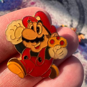 Puede incluir: Un colorido pin de esmalte con el personaje Mario de la serie de videojuegos Super Mario Bros. El pin representa a Mario con su clásico gorro rojo y peto, con un guante amarillo sosteniendo una seta. El pin mide aproximadamente 3,8 cm de altura.