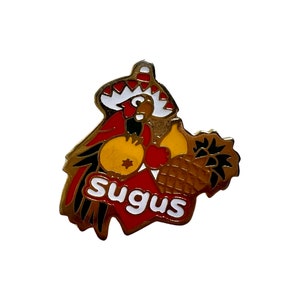 May include: A gold-coloured metal pin featuring a cartoon parrot wearing a red, white and yellow sombrero. The parrot is holding a pineapple and two yellow fruit. The word "Sugus" is written in white letters on a red background.