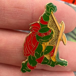 May include: A colourful enamel pin featuring a cartoon character in a fighting stance. The pin has a gold border and depicts a green figure with red accents, holding a yellow weapon. The character has a red mask.