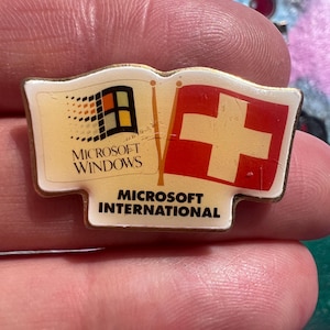 May include: A vintage Microsoft International pin featuring the Microsoft Windows logo and the Swiss flag. The pin has a gold-colored border and the text "MICROSOFT WINDOWS" and "MICROSOFT INTERNATIONAL".