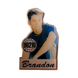 Puede incluir: Un pin vintage con un retrato de un hombre con una camisa azul. El pin incluye el texto "Beverly Hills 90210" en un diseño circular y el nombre "Brandon" debajo del retrato.