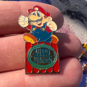 May include: A colourful enamel pin featuring the character Mario from the Super Mario Bros. video game. Mario is depicted in his classic outfit, jumping on a red and green rectangular base with text.