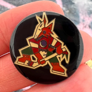 Könnte beinhalten: Runder, schwarzer Ansteckpin mit einem stilisierten Kojoten-Design in Rot, Grün und Beige. Der Kojote ist in einem geometrischen Stil dargestellt, mit einem roten und beigen Kopf und Körper. Der Pin ist wahrscheinlich ein Sammlerstück oder ein Souvenir.