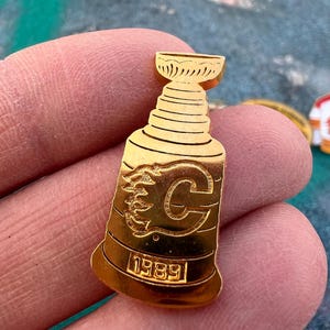 Könnte beinhalten: Goldfarbene Anstecknadel in Form eines Pokals. Die Nadel zeigt den Buchstaben "C" mit einem Flammenmuster und das Jahr "1989" am unteren Rand. Die Nadel wird zwischen zwei Fingern gehalten.