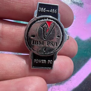 Może przedstawiać: Srebrna przypinka z napisem "IBM PS/1" i "POWER PC". Przypinka ma czarną obwódkę z numerami "386-486" na górze i czerwonym logo "V" pośrodku. Przypinka jest trzymana w dłoni.