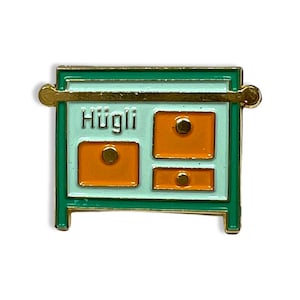 May include: A green and gold enamel pin shaped like a small dresser with three drawers. The top drawer has the word "Hyggli" written on it. The drawers are orange with gold knobs.