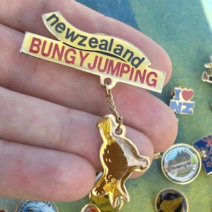 以下が含まれることがあります： 黒と赤の文字で「newzealand BUNGY JUMPING」と書かれたバナーが付いたゴールドトーンの金属製ピンバッジ。小さな金のチェーンがバナーと、金色のアクセントが入った茶色のキウイの形をしたぶら下がりチャームをつないでいます。