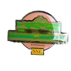 May include: A circular metal pin featuring a pink background with a green landscape illustration. A green train car is depicted with the text "BODENSEE-TOGGENBURG-BAHN" and "BISTROWAGEN" printed on it. A small yellow rectangular tab at the bottom displays "SSG".