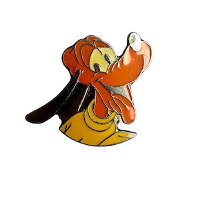 Puede incluir: Un pin de dibujos animados de Pluto, un perro marrón y naranja con una camisa amarilla, mirando hacia arriba con la lengua fuera.