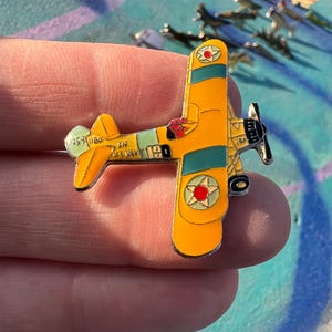 Puede incluir: Un pin de avión biplano amarillo con detalles verdes y rojos. El pin presenta una hélice, un piloto y el número "190". Las alas tienen rayas verdes y una estrella roja. El pin se sostiene entre los dedos.