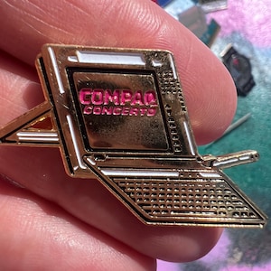 May include: Gold-coloured laptop pin with the text "COMPAQ CONCERTO" in pink on the screen. The pin features a keyboard design and a small stand. A vintage-style accessory.