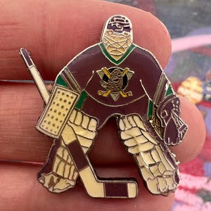 Könnte beinhalten: Ein Hockey-Torwart-Pin in den Farben Burgunder, Creme und Grün. Der Pin zeigt das Teamlogo und einen Torwart in voller Ausrüstung. Der Pin ist ein Sammlerstück.