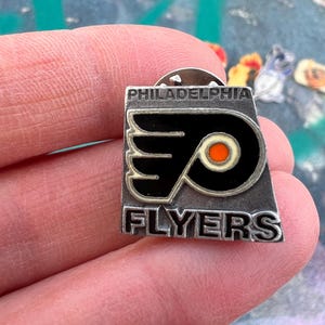 Könnte beinhalten: Ein silberfarbener Philadelphia Flyers-Anstecker. Der Anstecker zeigt das Logo des Teams in Schwarz, Orange und Weiß mit den Worten "PHILADELPHIA" und "FLYERS" in silberner Schrift. Der Anstecker ist rechteckig mit abgerundeten Ecken.