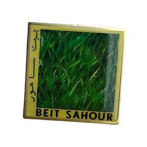 Può includere: Uno spillo in metallo dorato con un'immagine di campo verde e il testo "BEIT SAHOUR" in nero.