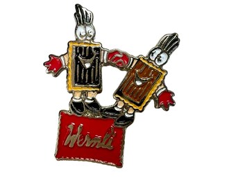 Könnte beinhalten: Eine goldfarbene Anstecknadel mit zwei Comicfiguren, die auf einem roten Kasten mit dem Aufdruck "Werther's" stehen. Die Figuren tragen braune und schwarze Outfits und haben rote Handschuhe.