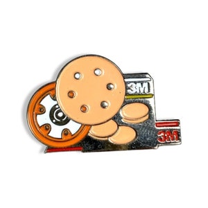 May include: A silver and orange enamel pin featuring a 3M logo and a depiction of sanding discs. The sanding discs are orange and have a white centre with a black circle in the centre. The pin is a great way to show your love for 3M products.
