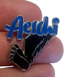 May include: A blue enamel pin with the word "Aerdi" in a stylized font. The pin features a black paw print with claws.