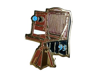 Puede incluir: Un pin de metal dorado de una cámara vintage con cuerpo marrón y detalles azules. La cámara está en un trípode y tiene el número "93" en el lateral.