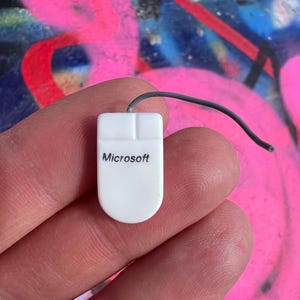 May include: A small, white Microsoft computer mouse with a black cord. The mouse has the Microsoft logo printed on it. The background is a colorful graffiti design with pink, red, and blue paint.