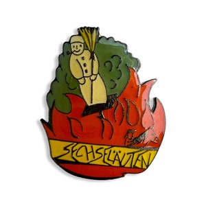 Peut inclure: Une épingle en émail rouge et jaune avec une figurine de dessin animé d'une personne tenant un balai debout devant un buisson vert. La figurine est en feu et le texte "SECHS LAUTEN" est sur une bannière jaune.