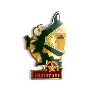 Pode incluir: Um alfinete dourado com um jacaré verde de desenho animado vestindo um colete estampado em amarelo e dourado. O jacaré está segurando um símbolo de balança dourado e uma estrela dourada. O alfinete tem uma faixa vermelha e dourada com o texto "Hürlimann" nela.