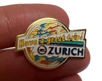 May include: A gold-toned pin with the words "Have a great trip!" and "ZURICH" in white and blue lettering. The pin features a colorful design of yellow, orange, and blue shapes, with a green and blue stripe. The pin is held by a person's fingers.
