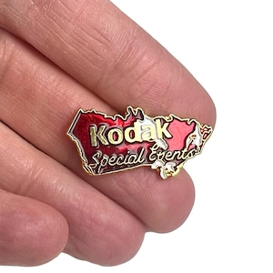 Op de afbeelding: Een gouden pin met een rode en witte omtrek in de vorm van de Verenigde Staten. De pin heeft de tekst "Kodak Special Events" in gouden letters.