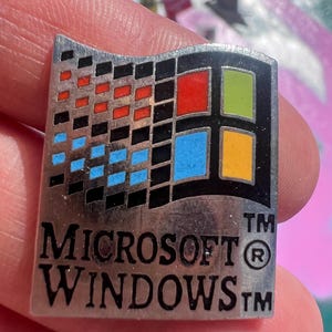 Puede incluir: Un pin plateado de Microsoft Windows con el logotipo clásico de Windows. El logotipo es un diseño de ventana de cuatro paneles con cuadrados rojos, verdes, azules y amarillos. El texto "MICROSOFT WINDOWS" está debajo.