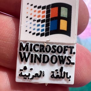 May include: A white rectangular pin featuring the Microsoft Windows logo. The logo includes a colourful four-pane window design and the words "MICROSOFT. WINDOWS." The pin also has Arabic text.