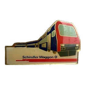 May include: A metal pin featuring a graphic of a red and blue train engine. The lower section is a cream-coloured rectangular base with the text Schindler Waggon and a small circular logo, underlined by three horizontal red lines in a gold frame.