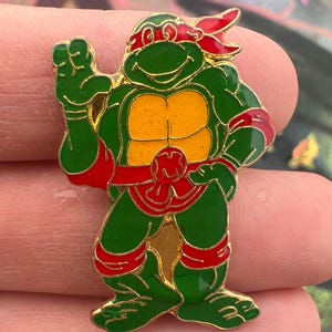 May include: A green and gold enamel pin featuring a cartoon turtle character with a red headband and accents. The pin has a gold border and a yellow chest. The turtle is in a standing pose with one arm raised.