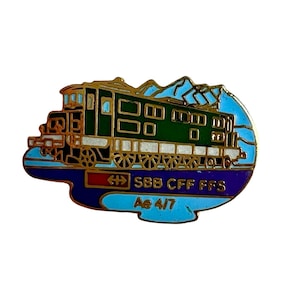 May include: A metal enamel pin featuring a green electric locomotive train against a light blue mountain backdrop. The pin includes the text SBB CFF FFS and Ae 4/7 in gold lettering on a dark blue and red background with gold trim detailing.