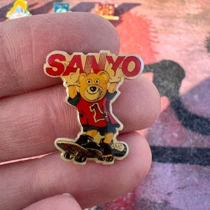 May include: A vintage Sanyo pin badge showing a cartoon bear on a skateboard. The bear is wearing a red jersey with the number 1 and black shorts. The word "SANYO" is in red above the bear.