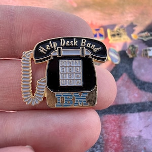 Könnte beinhalten: Ein Vintage-Pin in Form eines schwarzen Telefons mit goldenen Verzierungen. Der Pin zeigt den Text "Help Desk Bund" und das IBM-Logo in Blau. Das Telefon hat ein weißes Spiralkabel und eine Tastatur mit Zahlen.