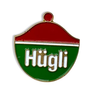 May include: A gold-colored pin with a red, white, and green design. The pin features the word "Hügli" in white letters with a gold outline.