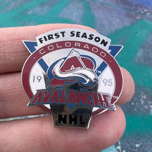 Könnte beinhalten: Metall-Anstecknadel mit dem Logo der Colorado Avalanche, mit dem Text "FIRST SEASON COLORADO 1995 AVALANCHE NHL". Die Anstecknadel hat ein Farbschema aus Rot, Weiß, Blau und Silber. Die Anstecknadel hat die Form eines Schildes.