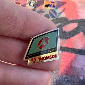 Può includere: Una spilla color oro con un design verde e rosso, con la scritta "THOMSON". La spilla ha un bordo nero e un logo stilizzato. Altre spille sono visibili sullo sfondo.