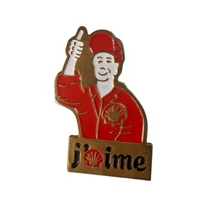 Puede incluir: Un pin de color dorado con una imagen de dibujos animados de un hombre con un mono rojo y un sombrero blanco. El hombre está haciendo un pulgar hacia arriba. El pin tiene el texto "j'aime" en la parte inferior.