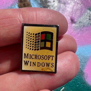 Könnte beinhalten: Eine rechteckige Anstecknadel mit dem Microsoft Windows-Logo. Die Anstecknadel hat einen gelben Hintergrund mit dem Windows-Logo in der oberen rechten Ecke und den Worten "MICROSOFT WINDOWS" in schwarzem Text darunter. Die Anstecknadel wird in einer Hand gehalten.