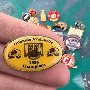 Könnte beinhalten: Eine Sammlung von Emaille-Anstecknadeln mit Logos und Emblemen von Hockeyteams. Die Hauptanstecknadel ist gelb und trägt den Text "Colorado Avalanche Western Conference 1996 Champions". Weitere Anstecknadeln zeigen ein Trikot, eine Bierflasche und einen Puck.